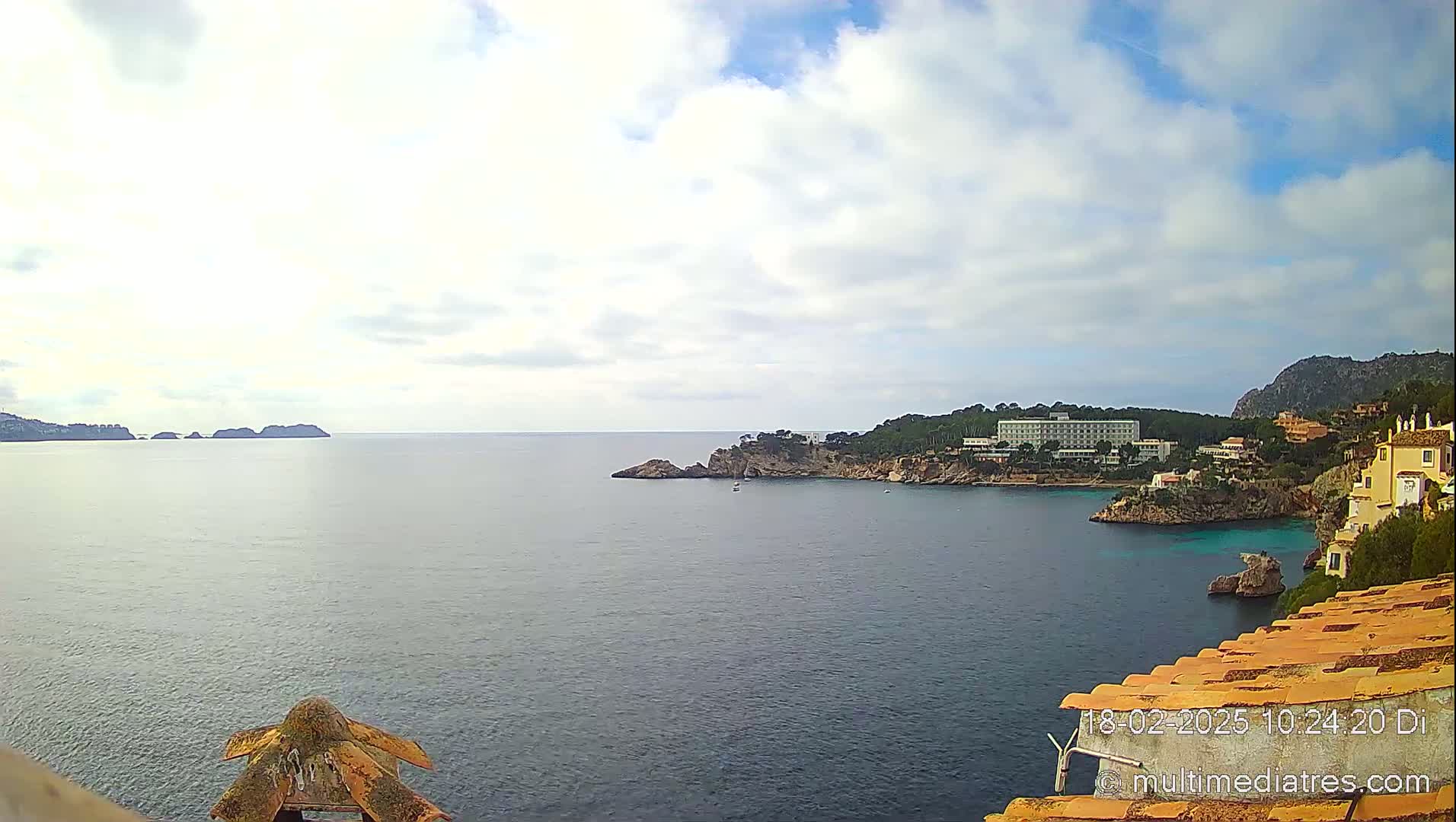 A partly cloudy day reveals a tranquil ocean scene with a coastline featuring buildings, trees, and rocky outcrops.