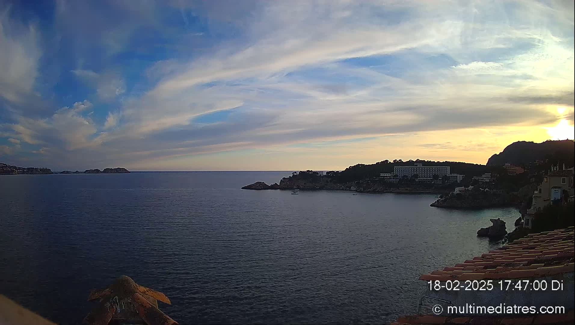 A tranquil coastal scene at sunset, featuring a calm sea, distant islands, a shoreline with buildings, and a partly cloudy sky.