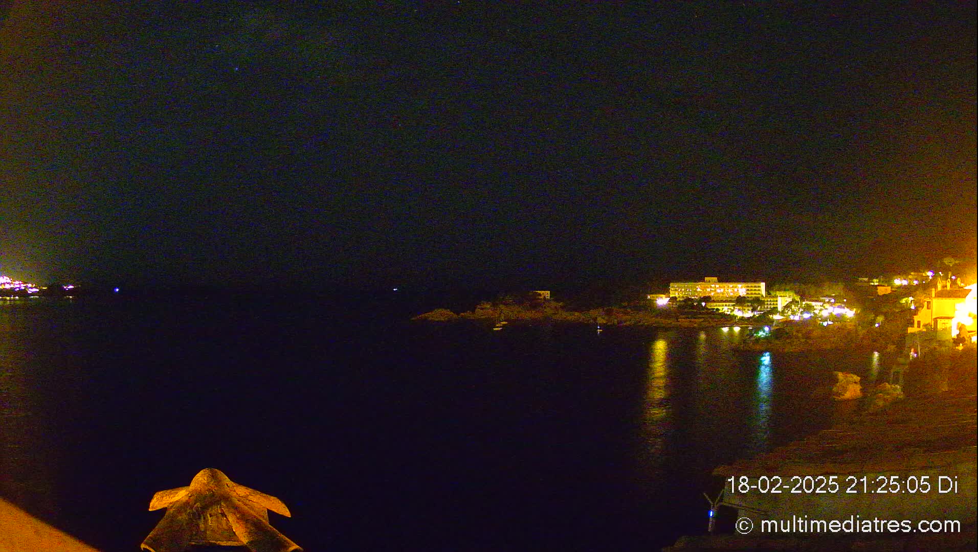 A nighttime view of a calm body of water reflecting the lights of buildings on the opposite shore under a clear, dark sky.