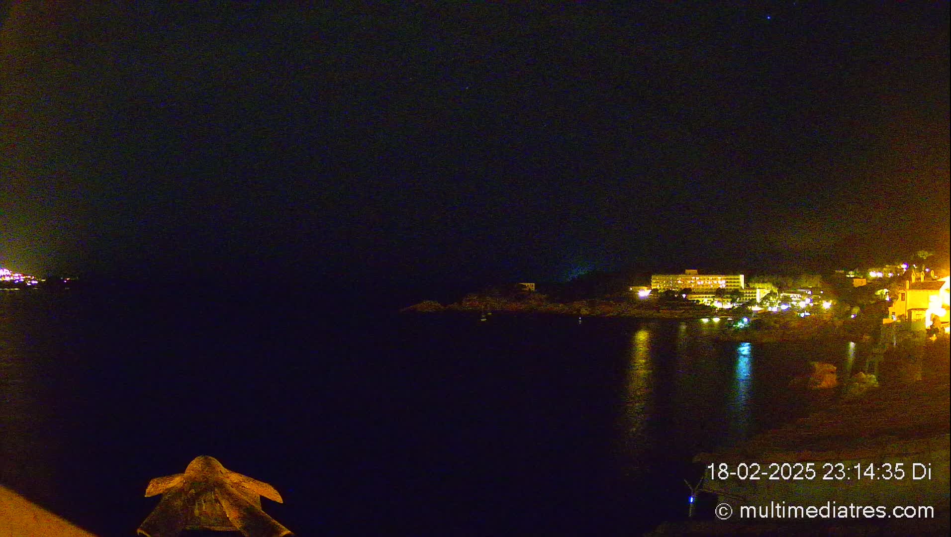 A nighttime view of a calm body of water reflecting the lights of buildings on the opposite shore under a clear, dark sky.