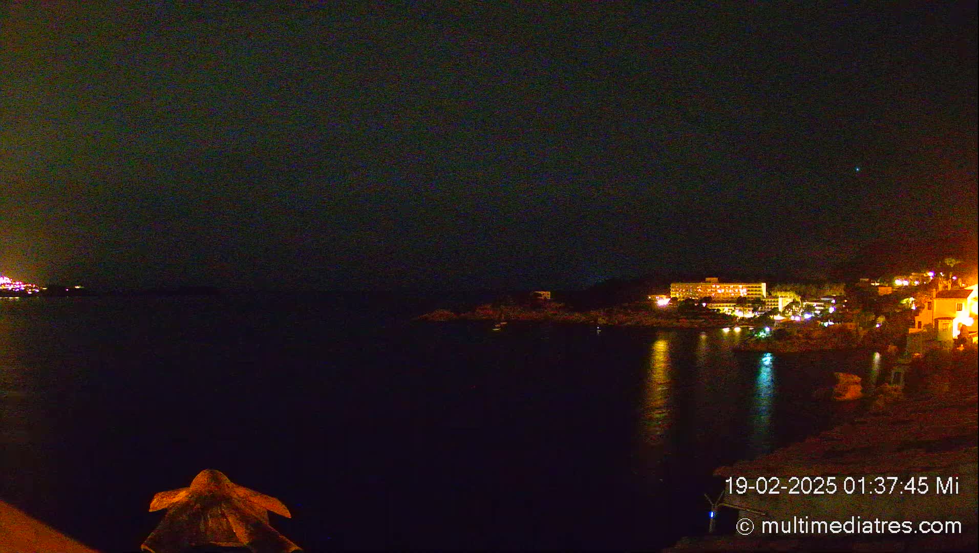 A nighttime view of a dark body of water reflecting lights from buildings on the opposite shore under a clear, dark sky.