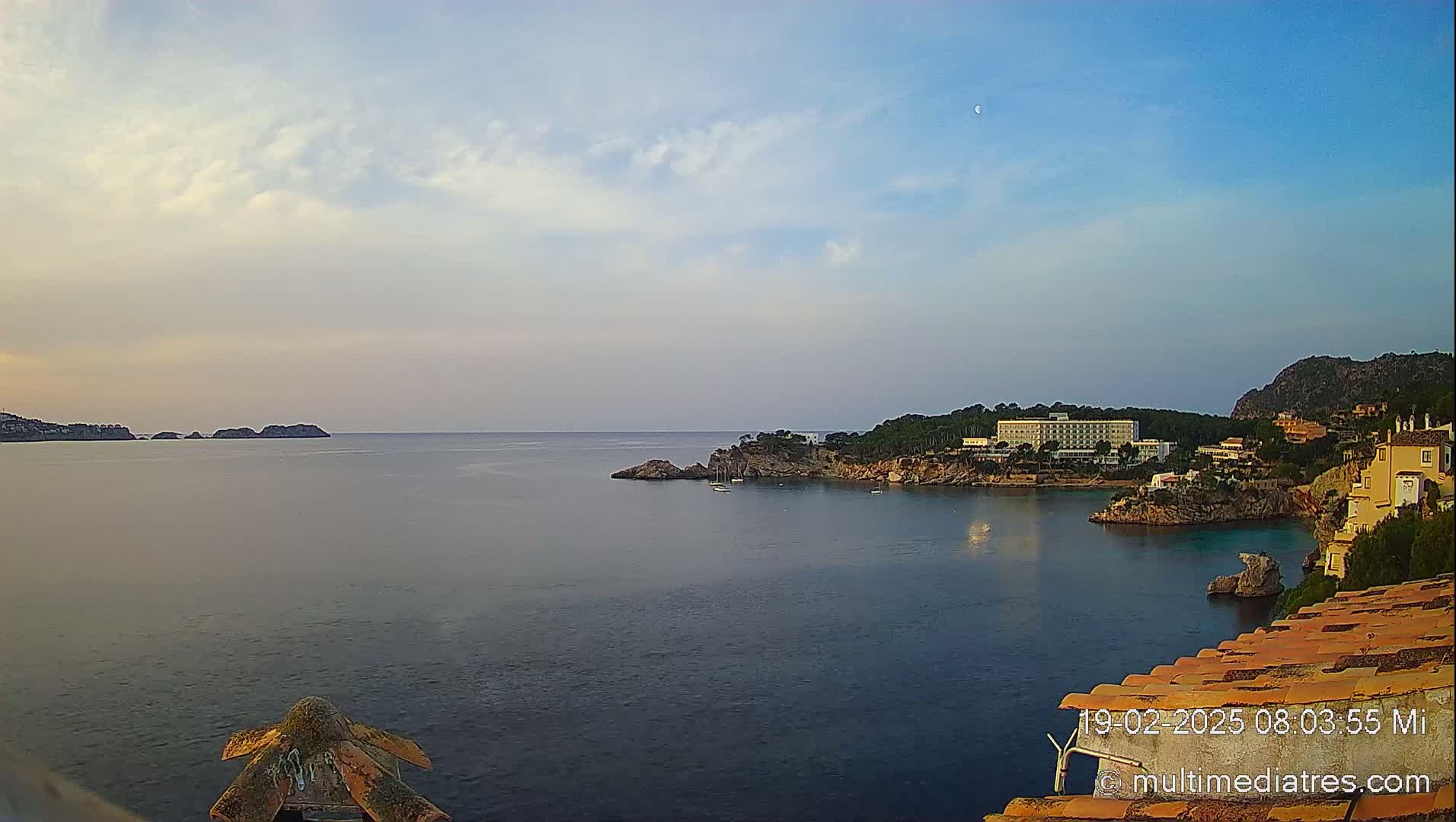 A calm, clear day shows a coastal scene with a large hotel and other buildings nestled into a rocky cove, a crescent moon visible in the sky, and several small boats in the water.