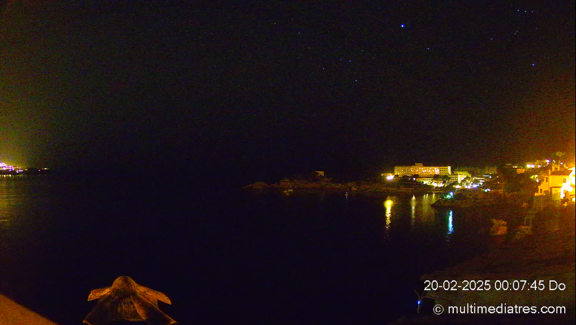 A nighttime view of a calm body of water reflecting the lights of buildings on the far shore under a clear, starry sky.