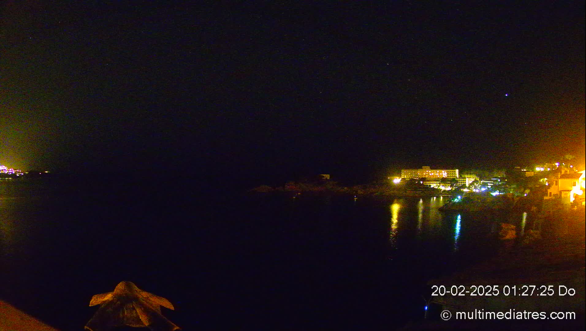 A nighttime view of a calm body of water reflecting the lights of buildings on the opposite shore under a clear, starry sky.