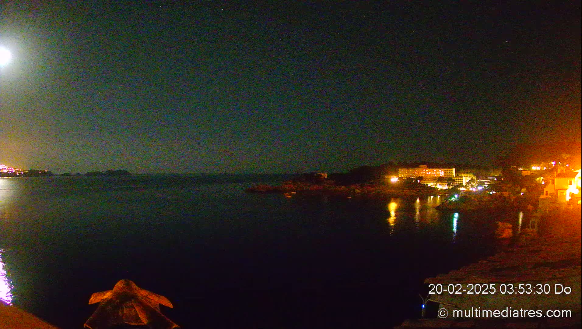A nighttime view of a calm body of water with a brightly lit hotel and other buildings on the shore under a clear, starry sky illuminated by a bright moon.