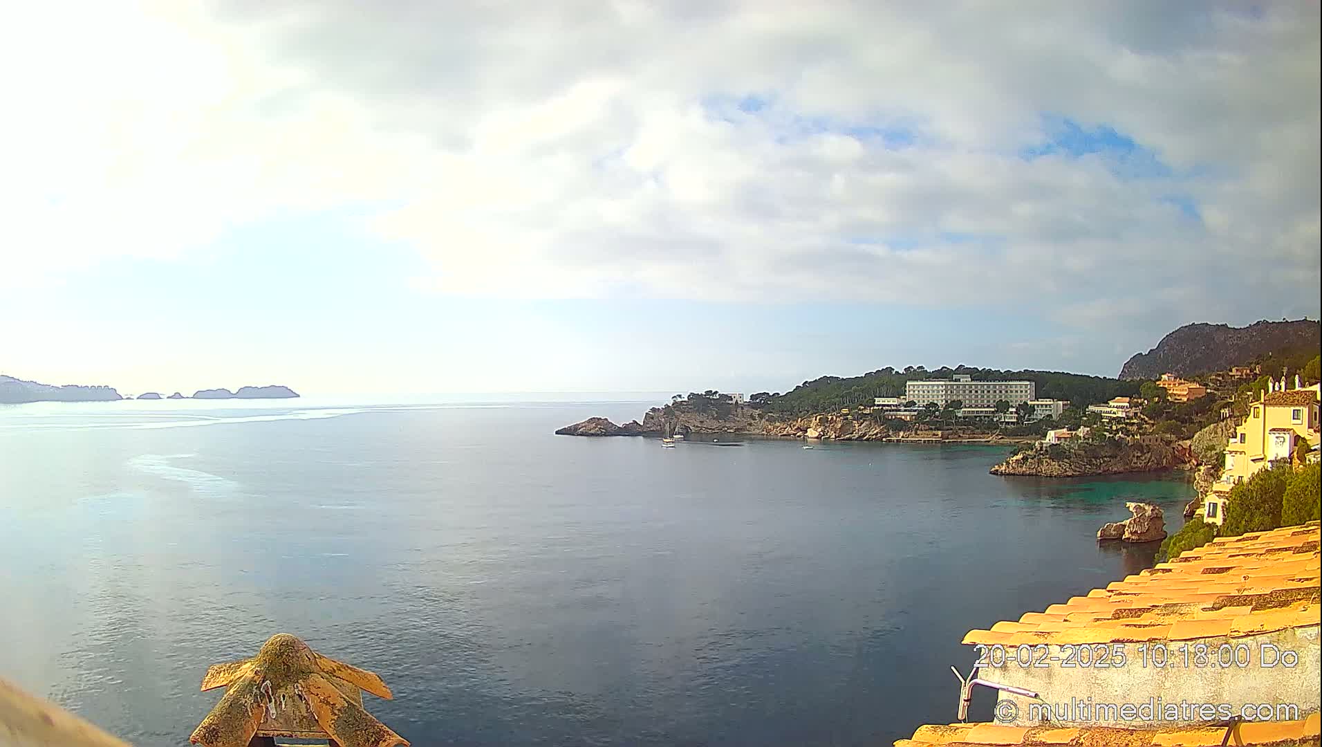 A partly cloudy day reveals a coastal scene with a calm sea, rocky inlets, a large hotel, and other buildings nestled along the shore.