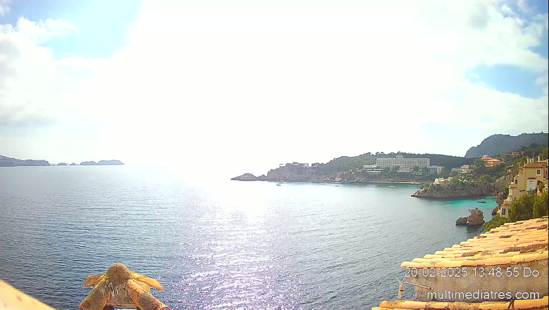 A sunny, partly cloudy day overlooks a calm ocean bay with distant islands, a shoreline featuring buildings and cliffs, and a tiled roof in the foreground.