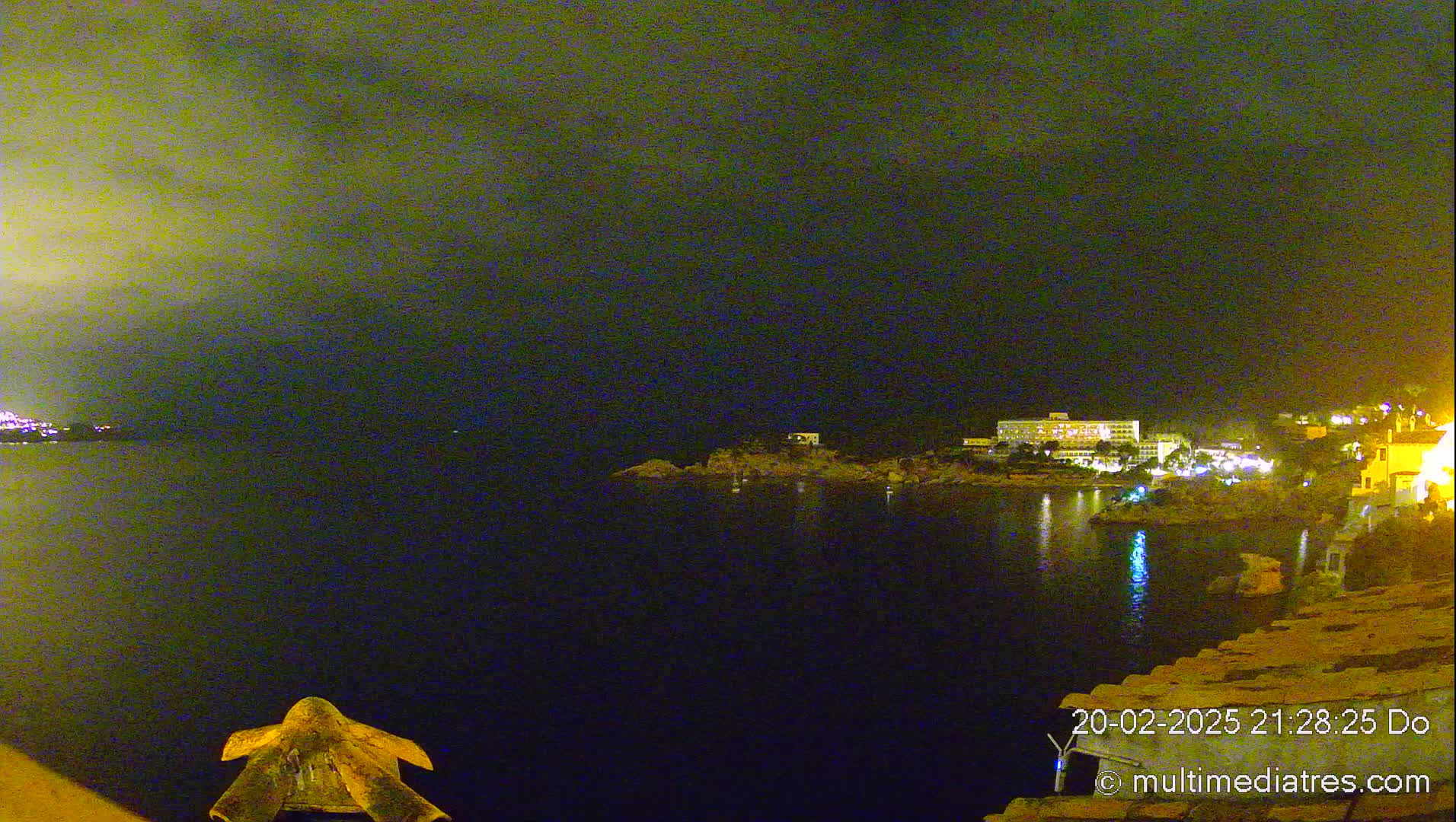A nighttime view of a dark body of water with a faintly lit shoreline featuring buildings and a large hotel across the water under a cloudy sky.