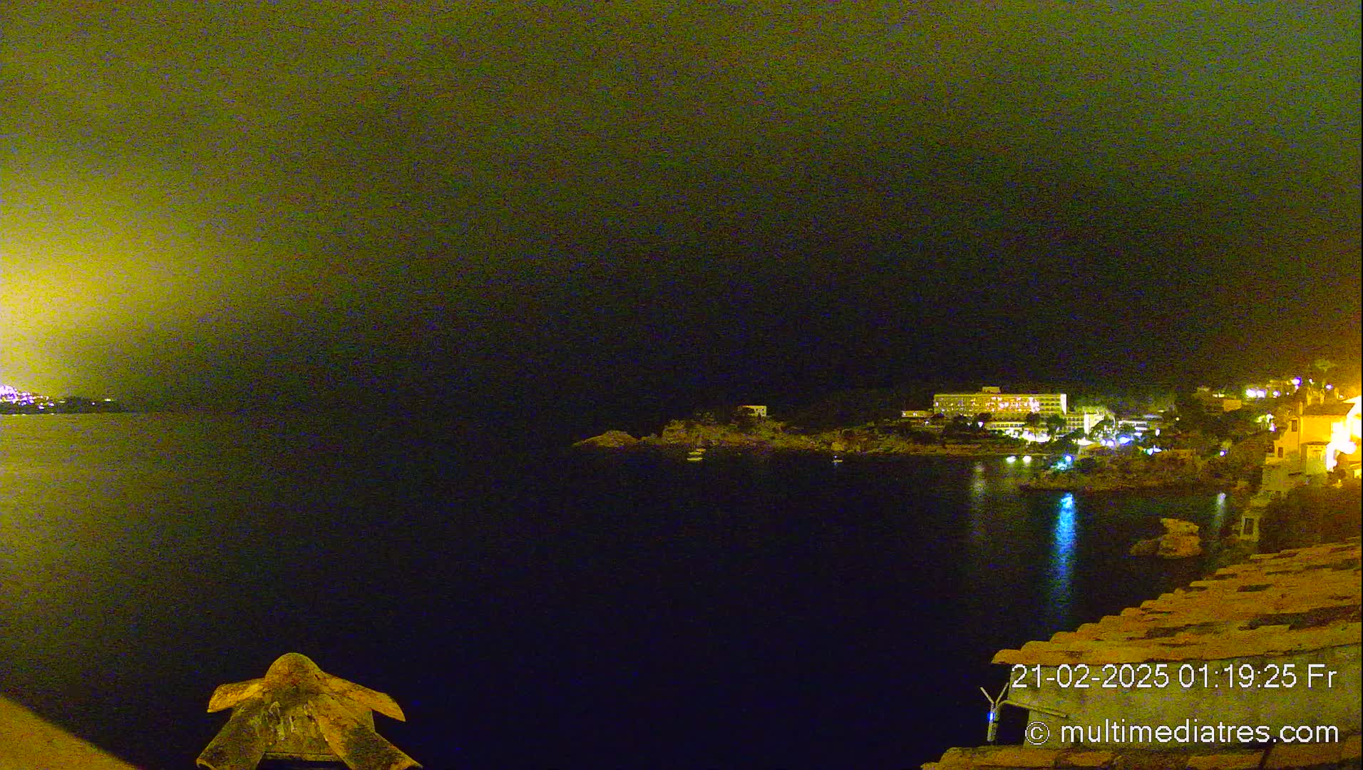 A nighttime coastal scene shows a dark body of water with a dimly lit shoreline featuring buildings and vegetation under a hazy, dark yellow-green sky.