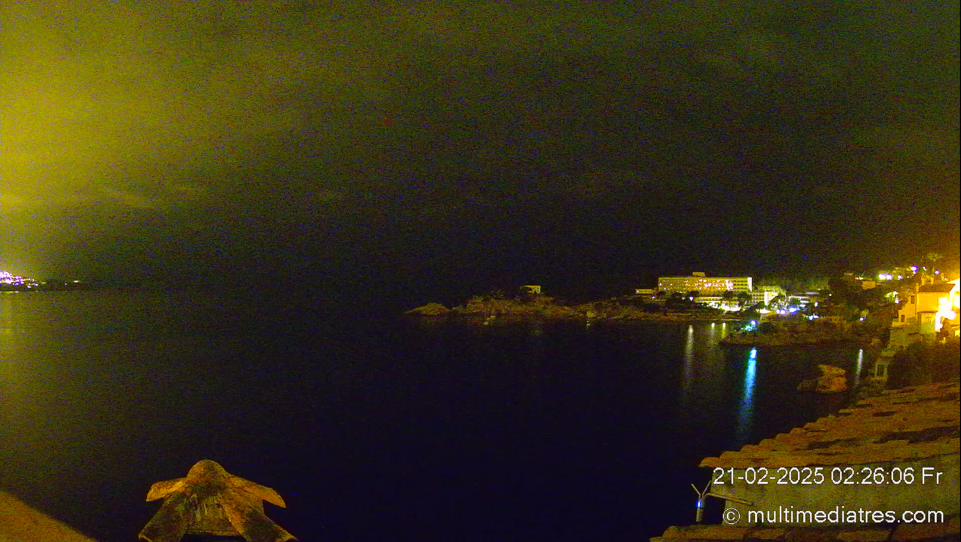 A nighttime view of a calm body of water with dimly lit buildings and vegetation on the opposite shore under a dark, yellow-tinged sky.