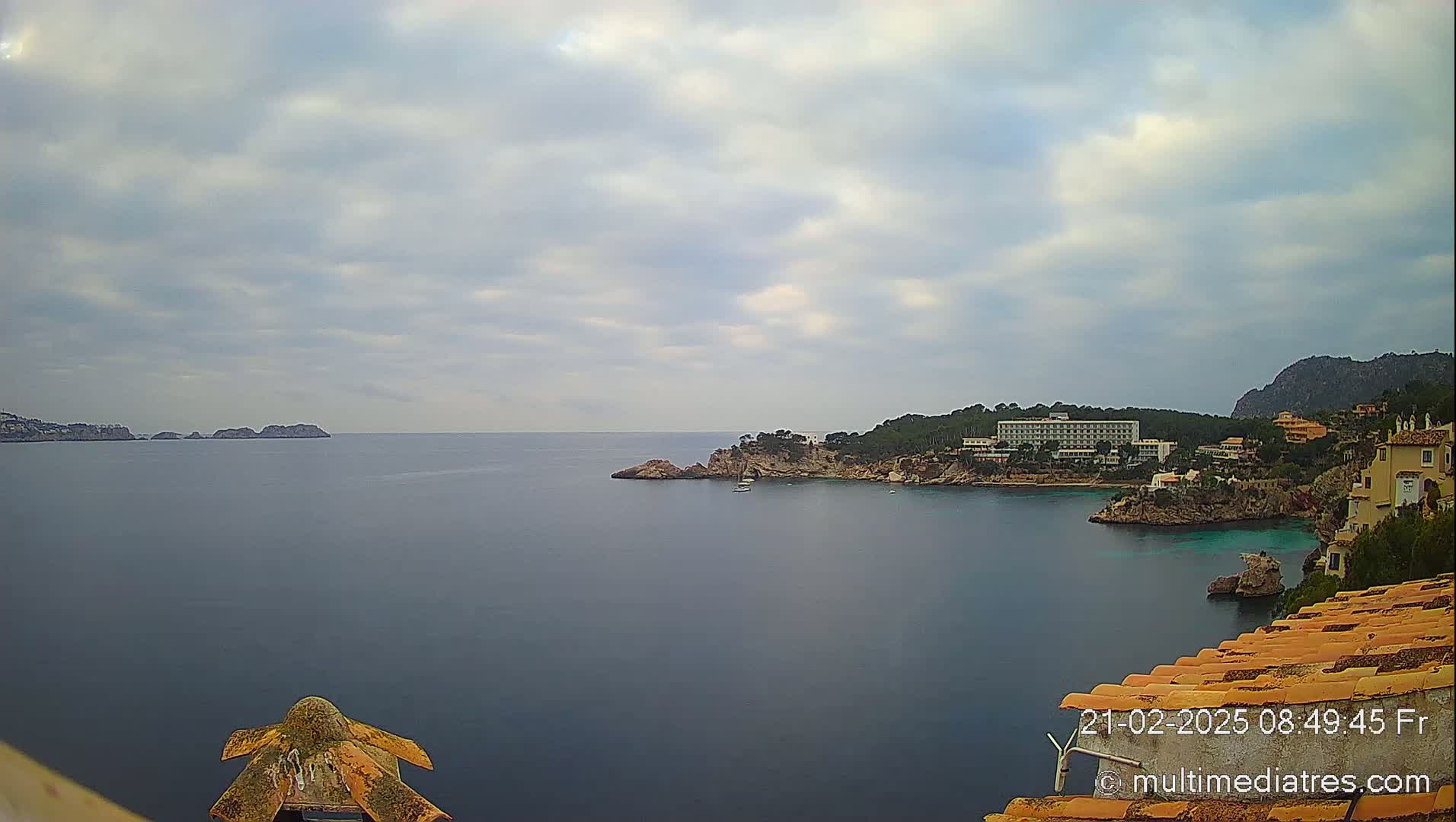 A tranquil ocean cove is seen under a cloudy sky, featuring a hotel and other buildings nestled on a rocky shoreline with small islands in the distance.