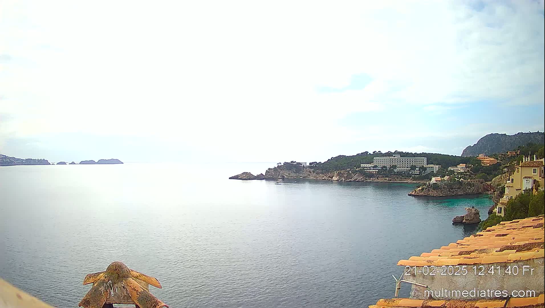 A calm sea stretches out to distant islands under a mostly cloudy sky, with a coastline featuring buildings and vegetation in the foreground.