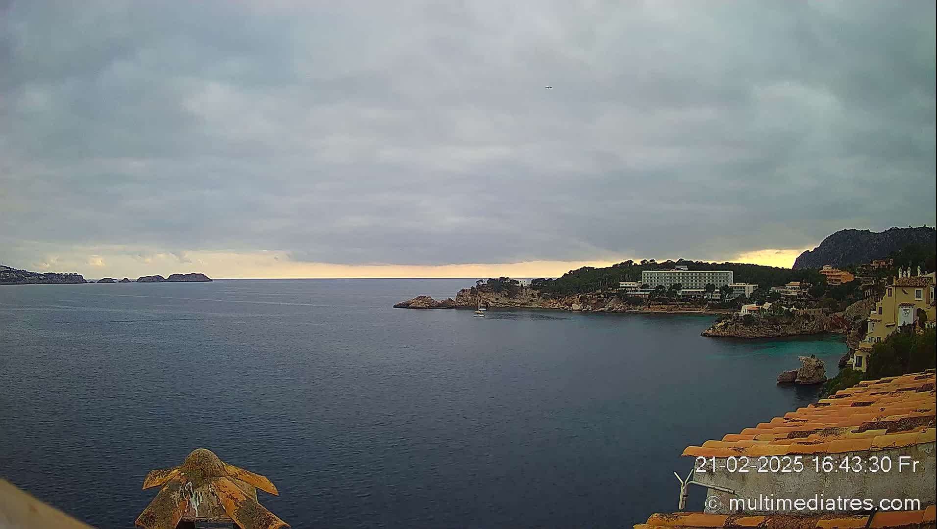 A coastal scene under a cloudy sky shows a calm sea with rocky headlands and buildings along the shoreline.