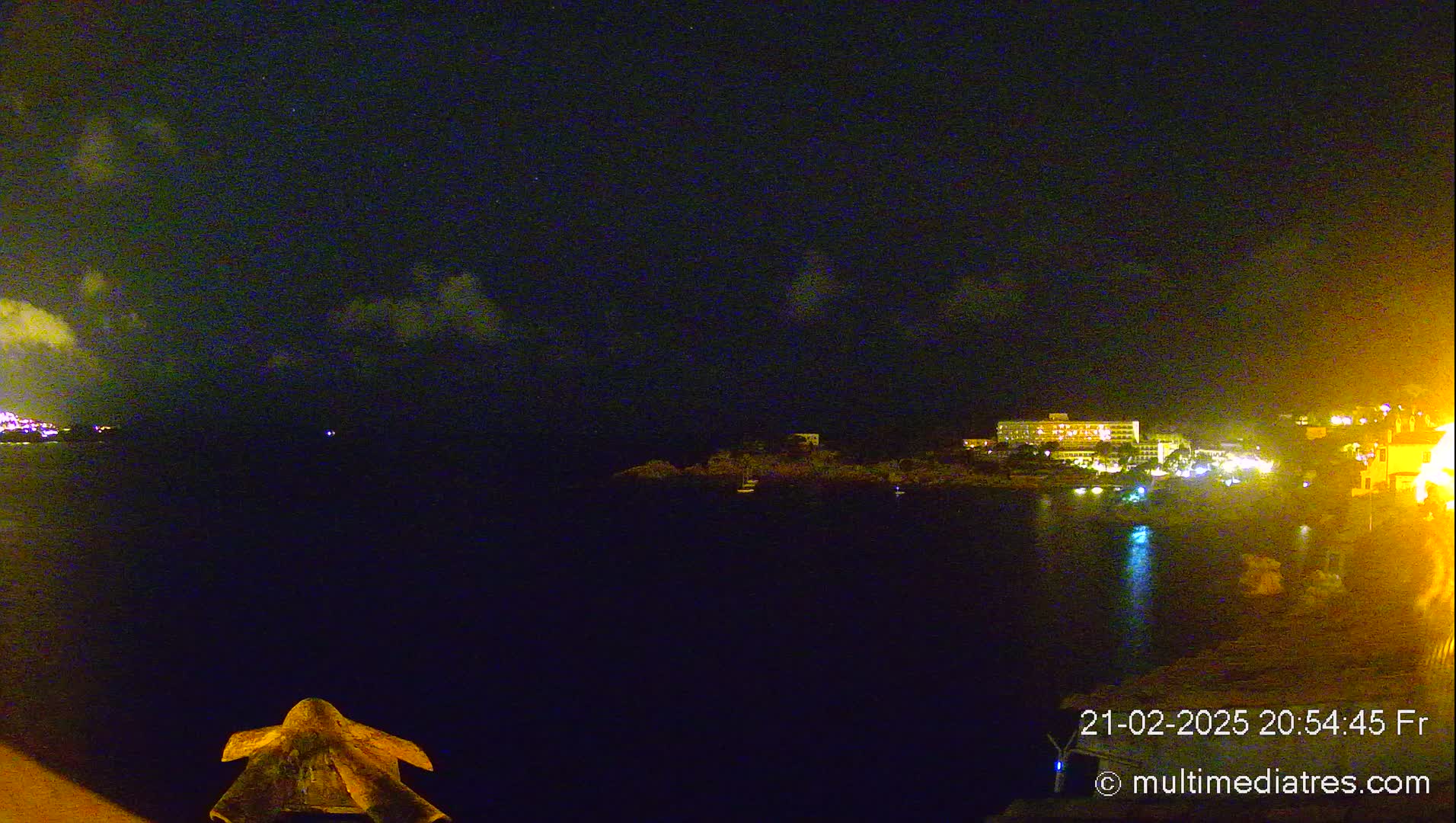 A nighttime coastal scene shows a dark body of water reflecting the lights of buildings on the opposite shore under a partly cloudy sky.
