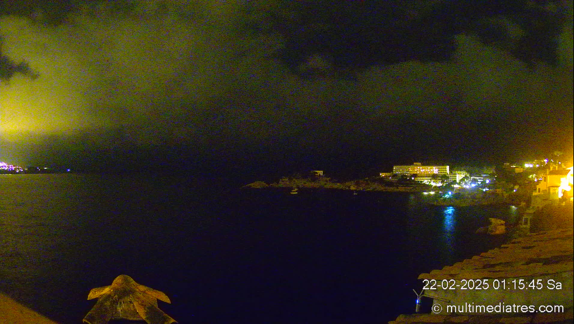 A nighttime coastal scene shows a dark body of water with a lit-up shoreline featuring buildings and a cloudy, dark sky.