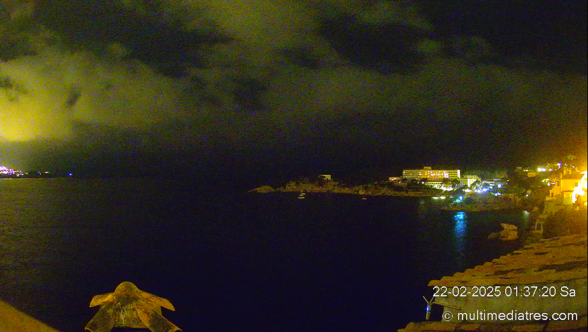 A nighttime view of a calm, dark body of water with illuminated buildings and land on the far shore under a cloudy sky.
