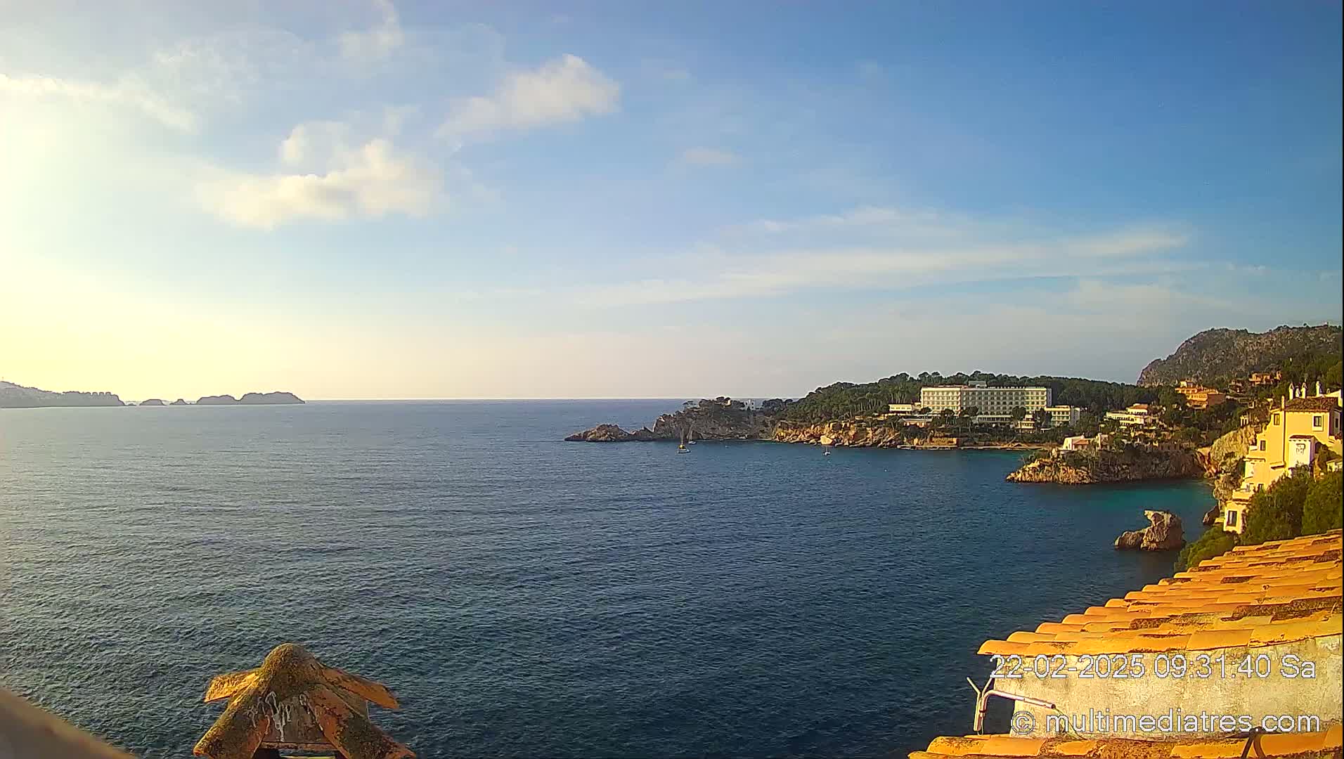 A calm, blue sea stretches to a distant coastline featuring a large hotel and other buildings nestled among trees and cliffs under a mostly clear sky.