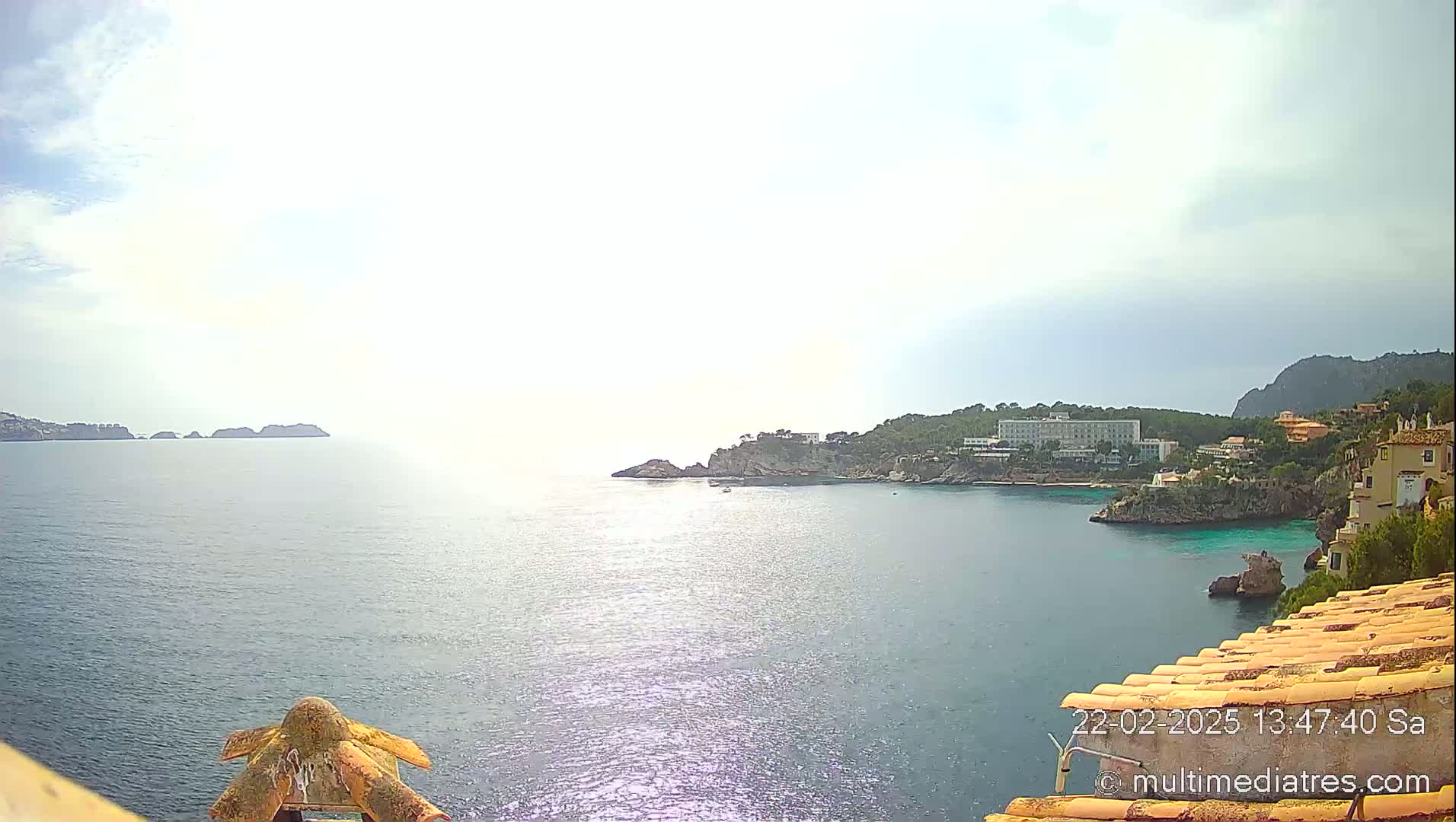 A partly cloudy day reveals a coastal scene with calm blue water, a rocky shoreline featuring buildings and vegetation, and distant islands under a bright sky.