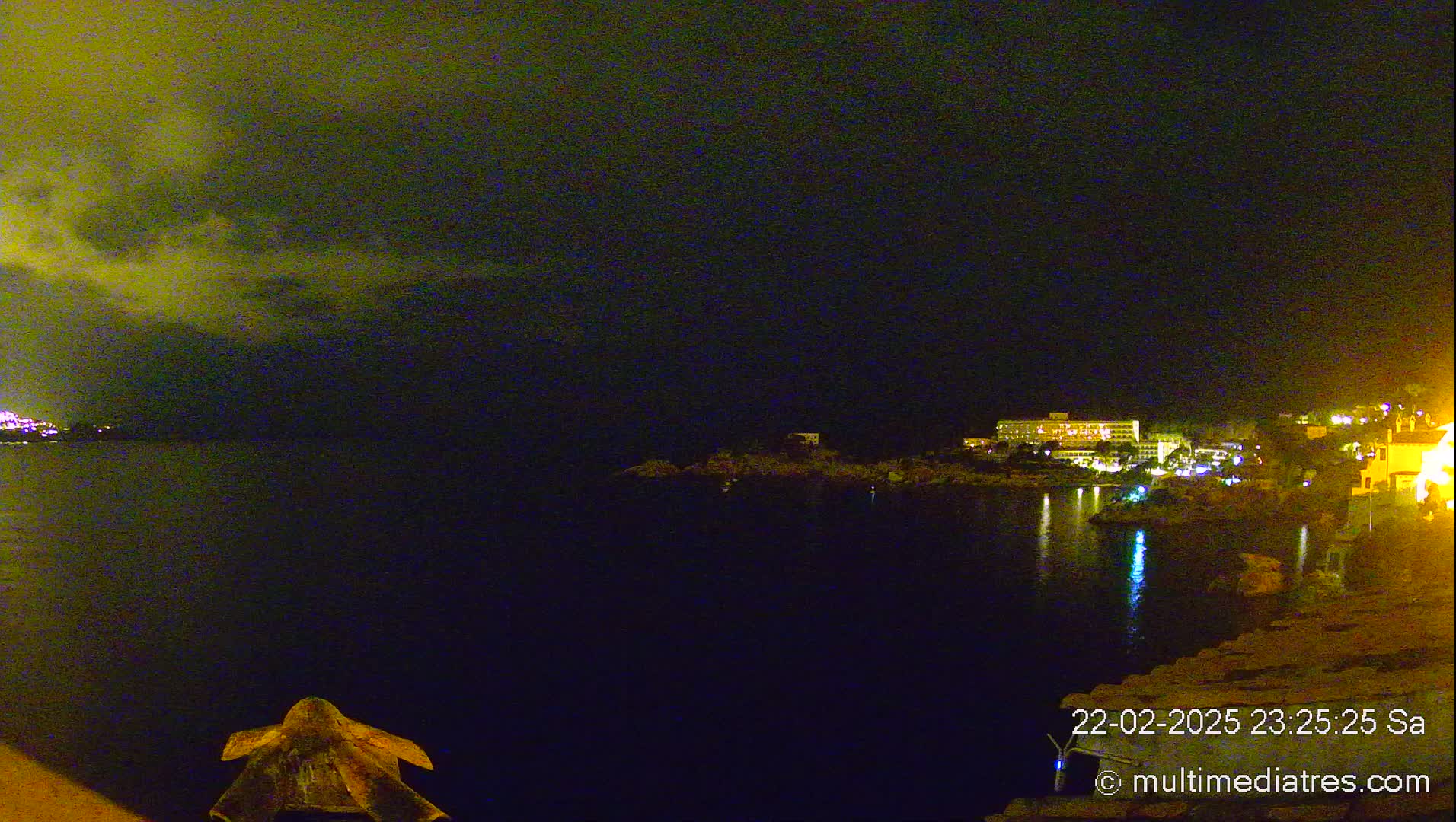 A nighttime view of a calm body of water with a dimly lit shoreline featuring buildings and a cloudy sky.