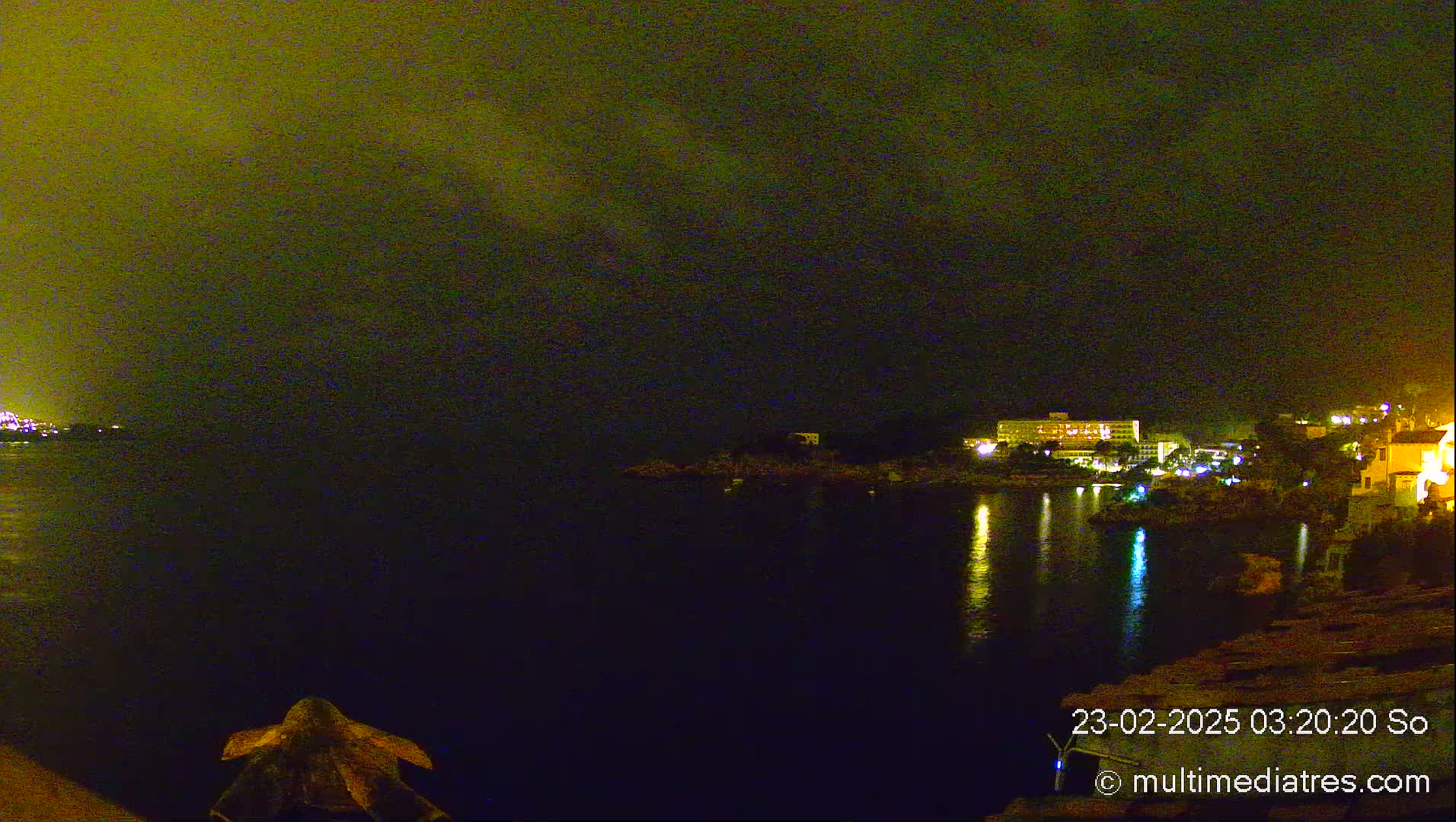 A nighttime view of a calm body of water with illuminated buildings and trees on the opposite shore under a dark, cloudy sky.