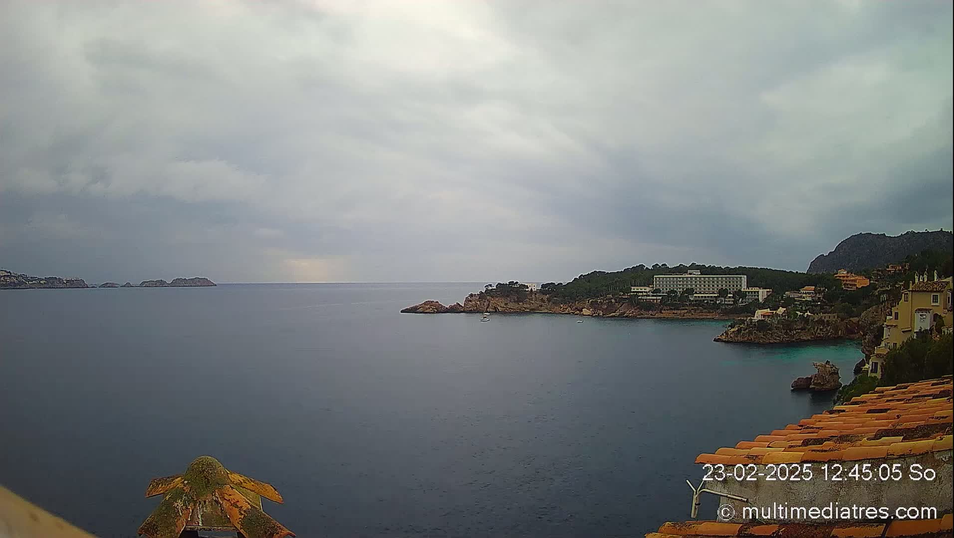 A calm, overcast day reveals a coastal scene with a hotel, rocky shoreline, and several small islands in the distance.