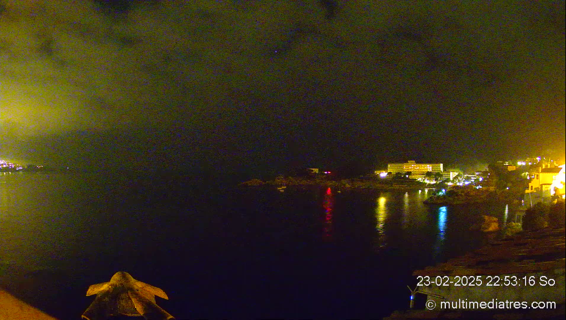 A nighttime view of a calm body of water with illuminated buildings and land on the opposite shore under a dark, cloudy sky.
