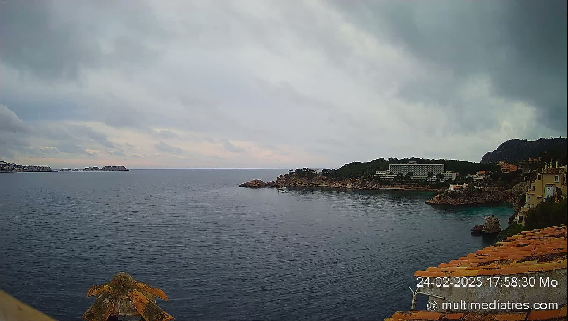 A calm, overcast day shows a coastal scene with a body of water, distant islands, a shoreline with buildings and trees, and an orange tiled roof in the foreground.