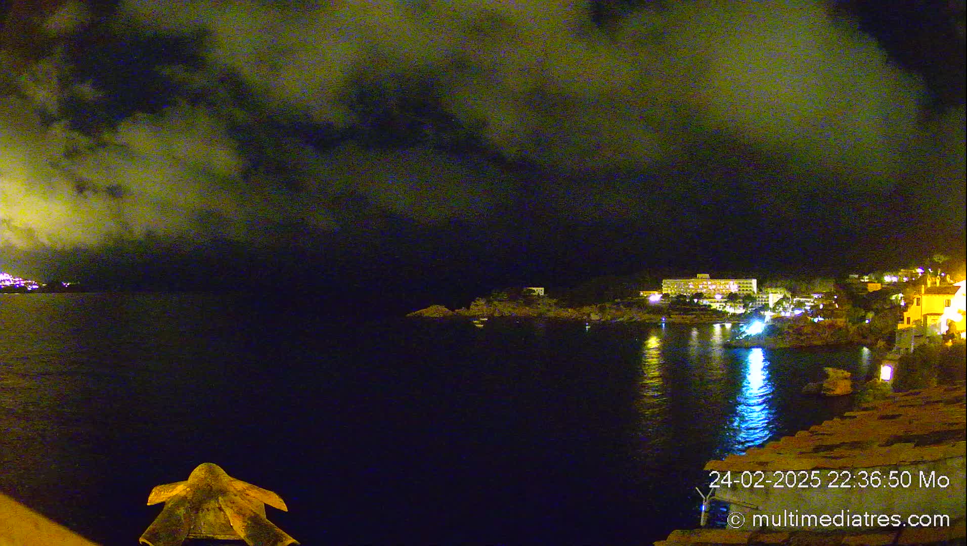 A nighttime view of a dark, calm body of water with a city's lights reflecting on its surface, under a cloudy sky.