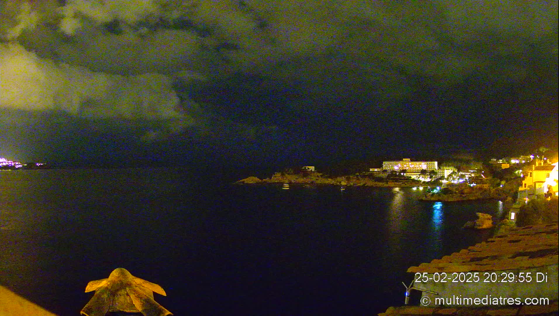 A nighttime coastal scene shows a dark body of water reflecting the lights of buildings on the shore under a cloudy sky.