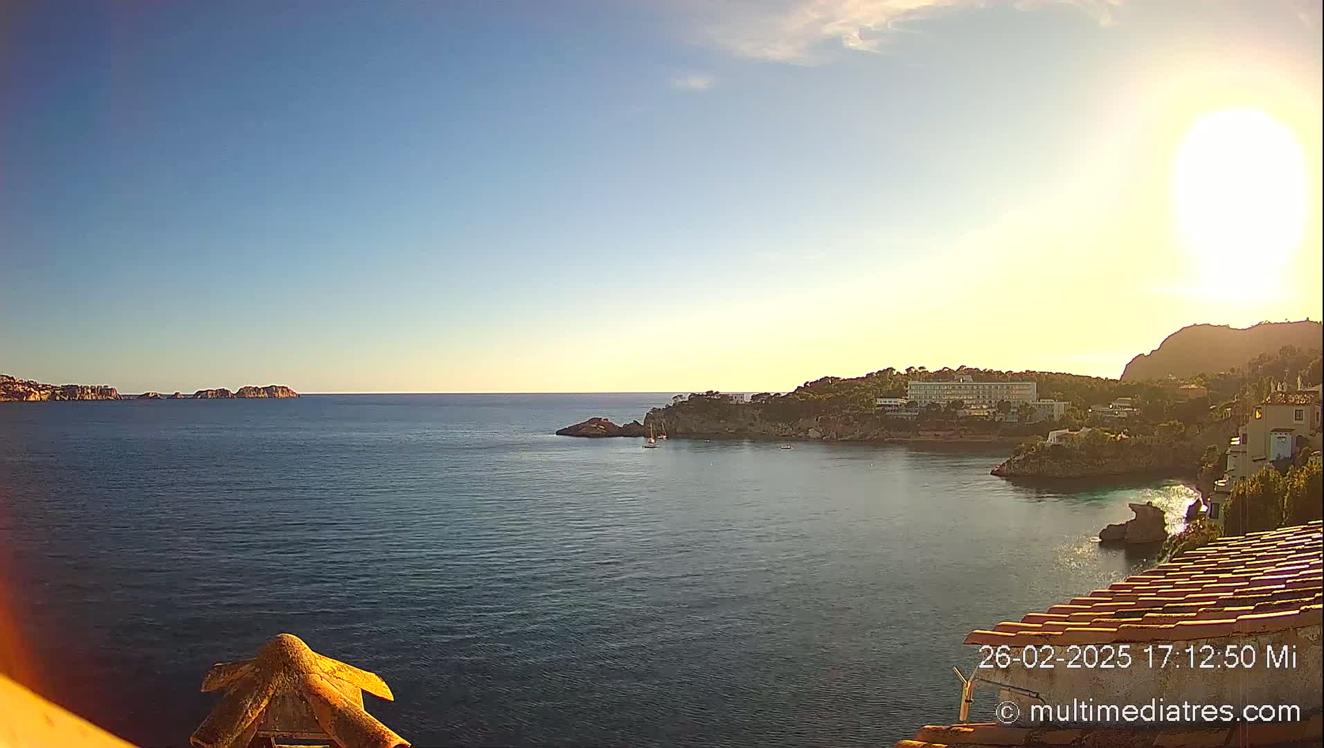 A tranquil ocean cove is seen under a mostly sunny sky, with a small sailboat visible in the distance near a rocky shoreline and buildings.