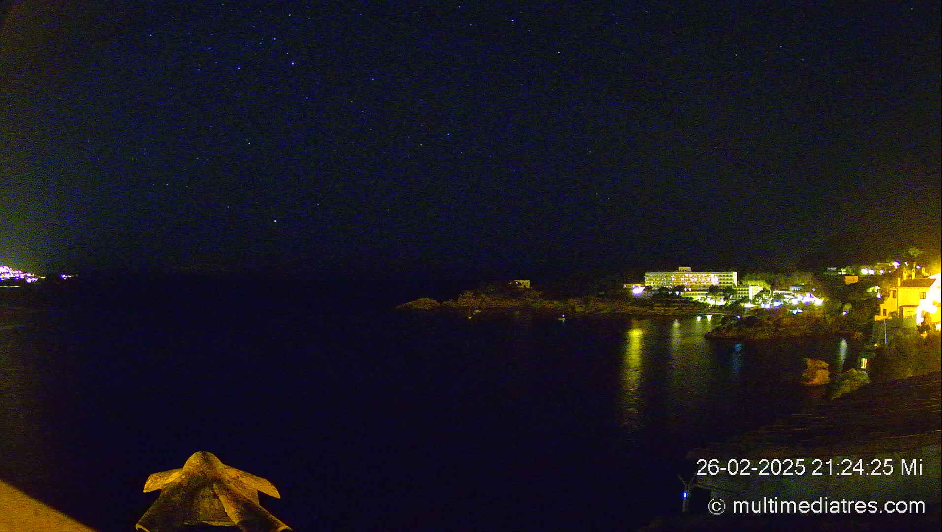 A nighttime view of a calm body of water with a building and other lights illuminating the shoreline under a clear, starry sky.