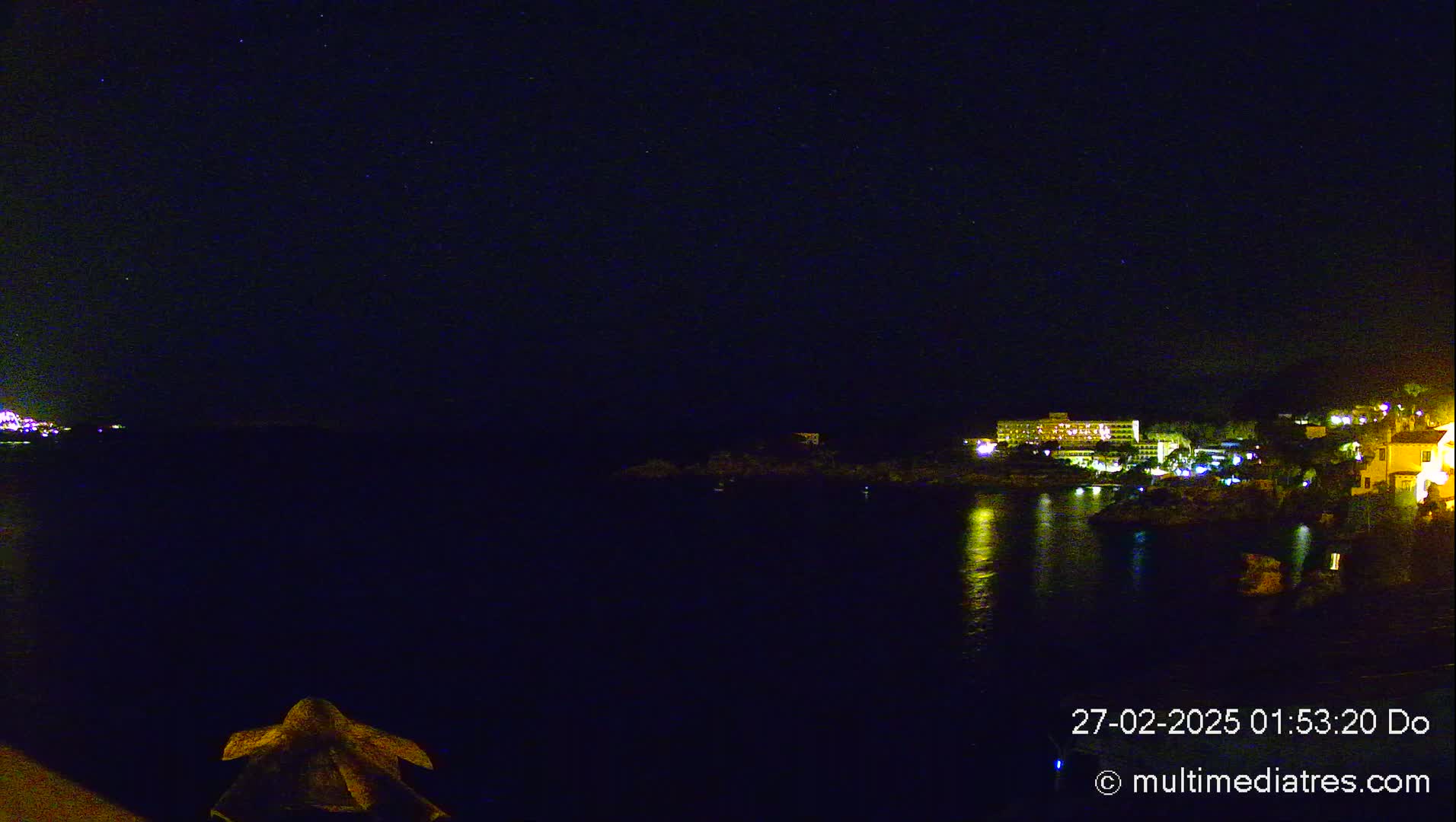A dark, nighttime coastal scene shows a calm body of water reflecting the lights of buildings on the opposite shore under a clear, starry sky.