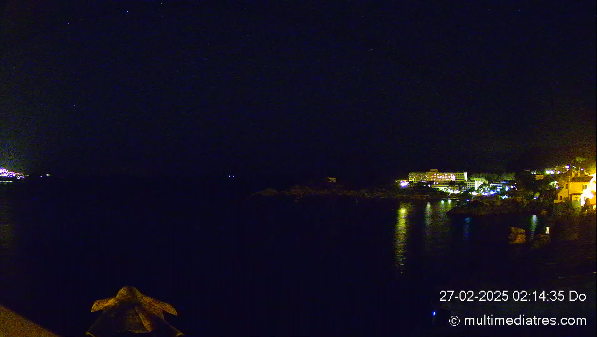 A nighttime view of a calm body of water reflecting the lights of buildings on the shore under a clear, starry sky.