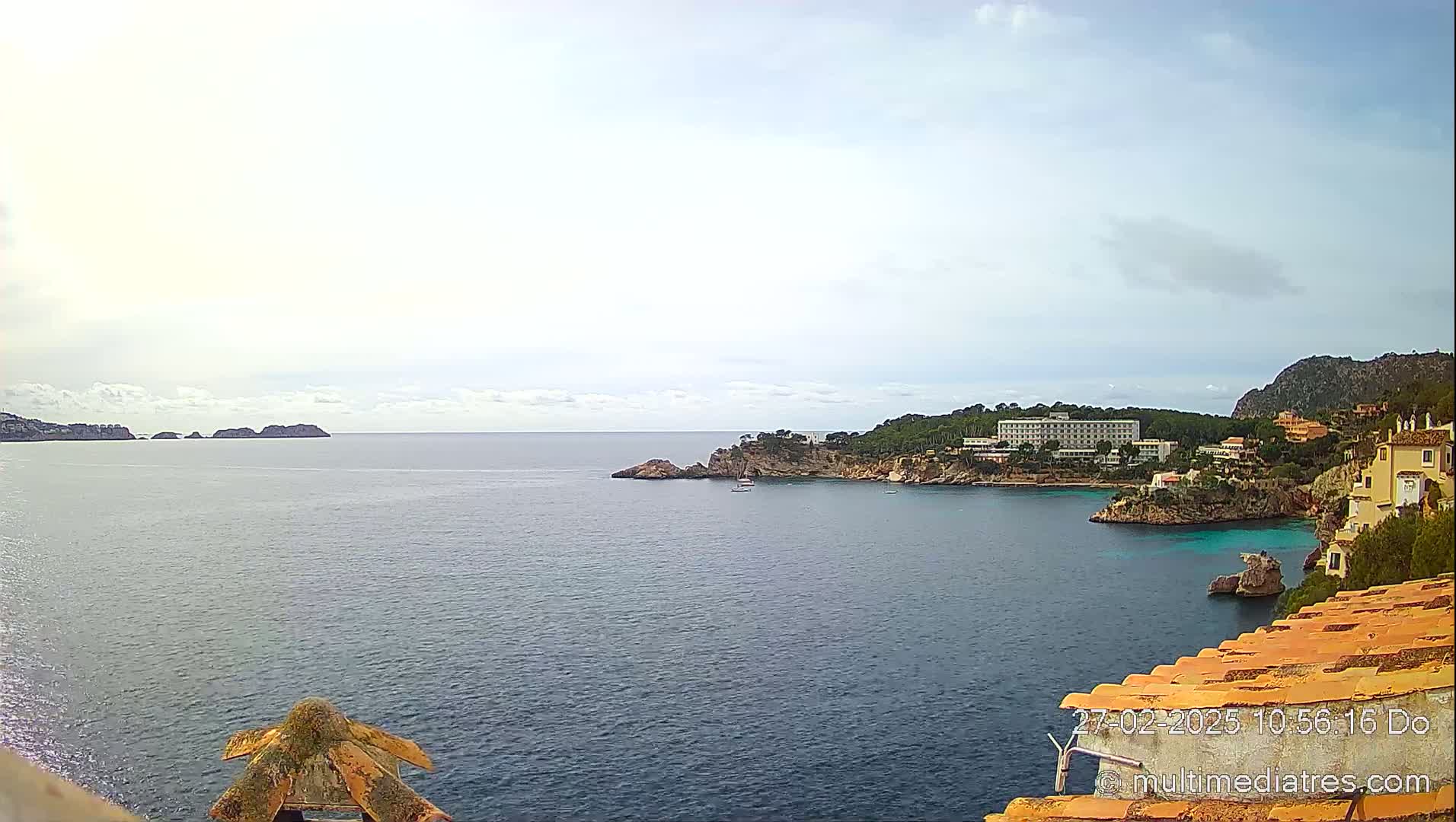 A partly cloudy day reveals a coastal scene of calm, dark-blue water, small islands in the distance, a hotel nestled amongst greenery on a rocky shore, and terracotta rooftops in the foreground.
