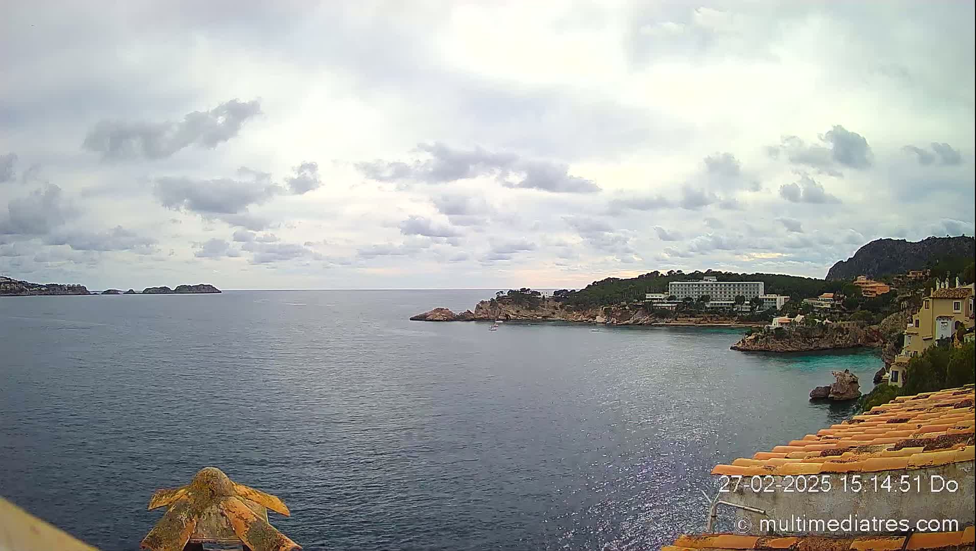 A coastal scene under a cloudy sky shows a calm sea with rocky inlets, a large hotel, and other buildings nestled along the shoreline.