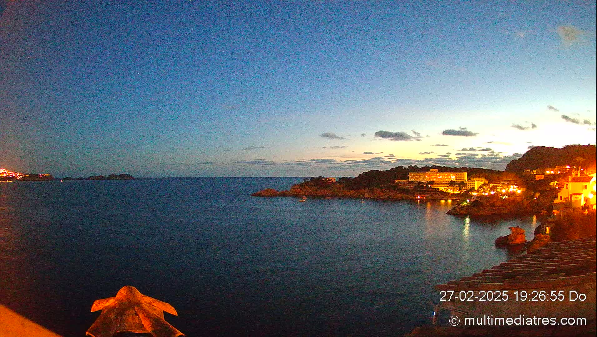A twilight view of a dark-blue sea with a coastline featuring illuminated buildings and a rocky shore under a mostly clear, dusky sky.
