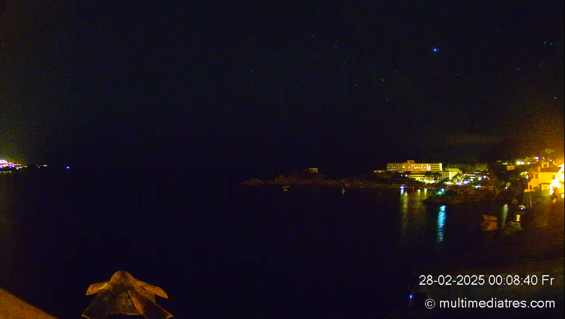 A nighttime coastal scene shows a dark body of water reflecting the lights of buildings on the opposite shore under a clear, starlit sky.