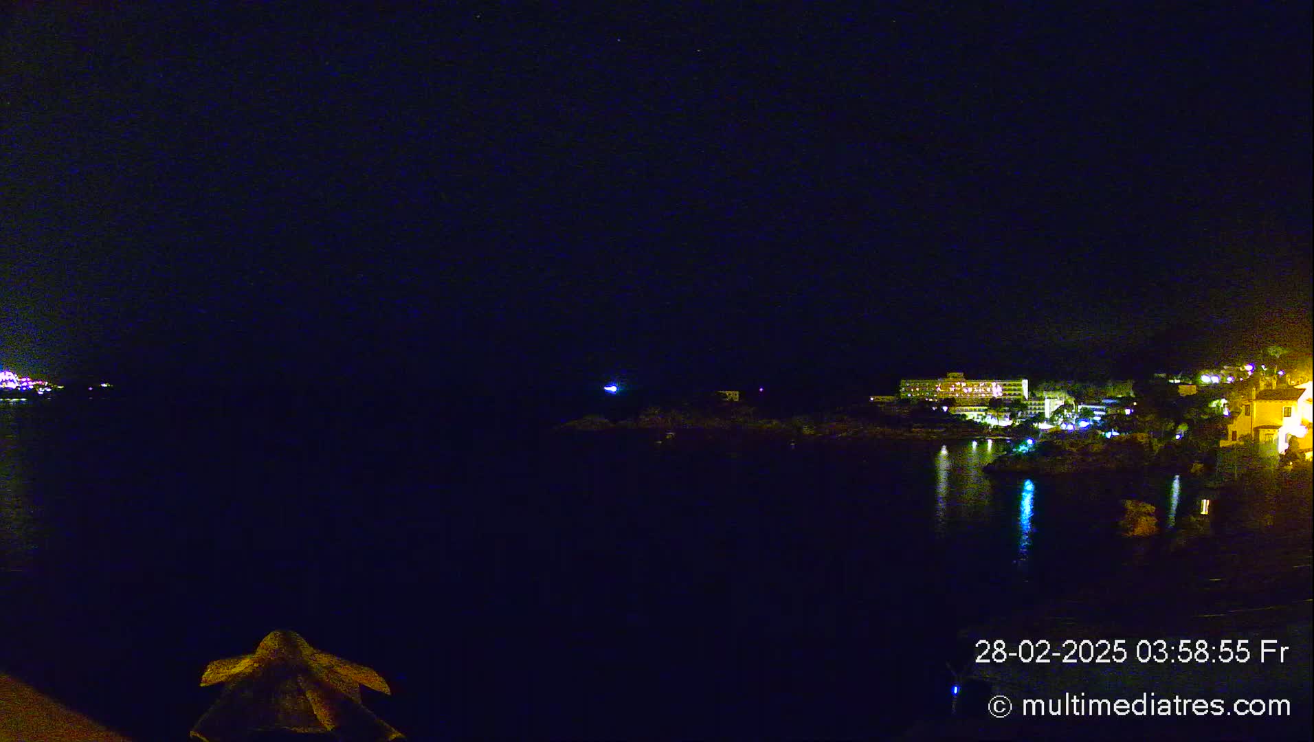 A nighttime view of a calm body of water reflecting the lights of buildings on the opposite shore under a clear, dark sky.