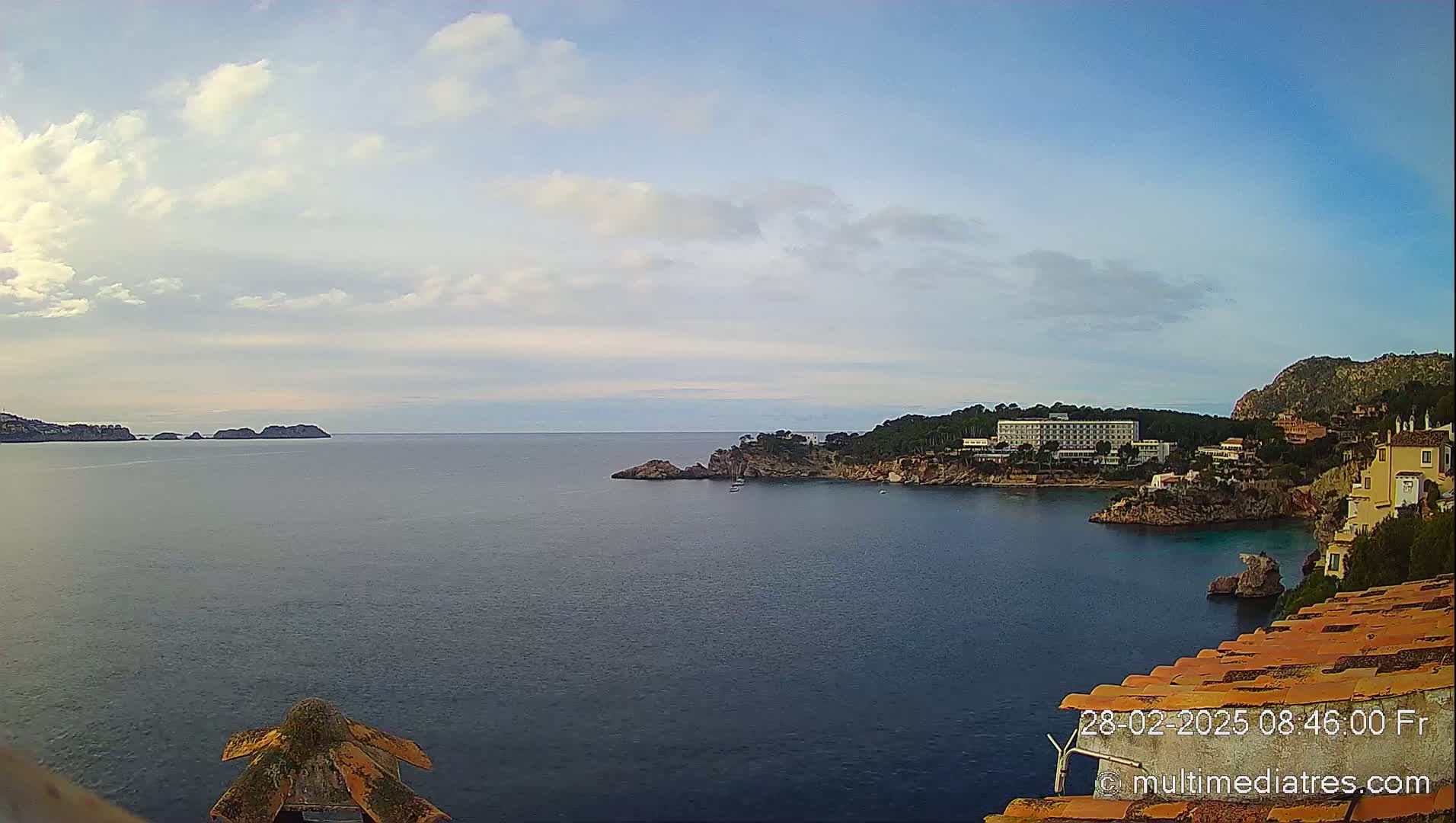 A partly cloudy sky overlooks a calm sea with a coastline featuring a large hotel and other buildings nestled among rocky cliffs and vegetation.