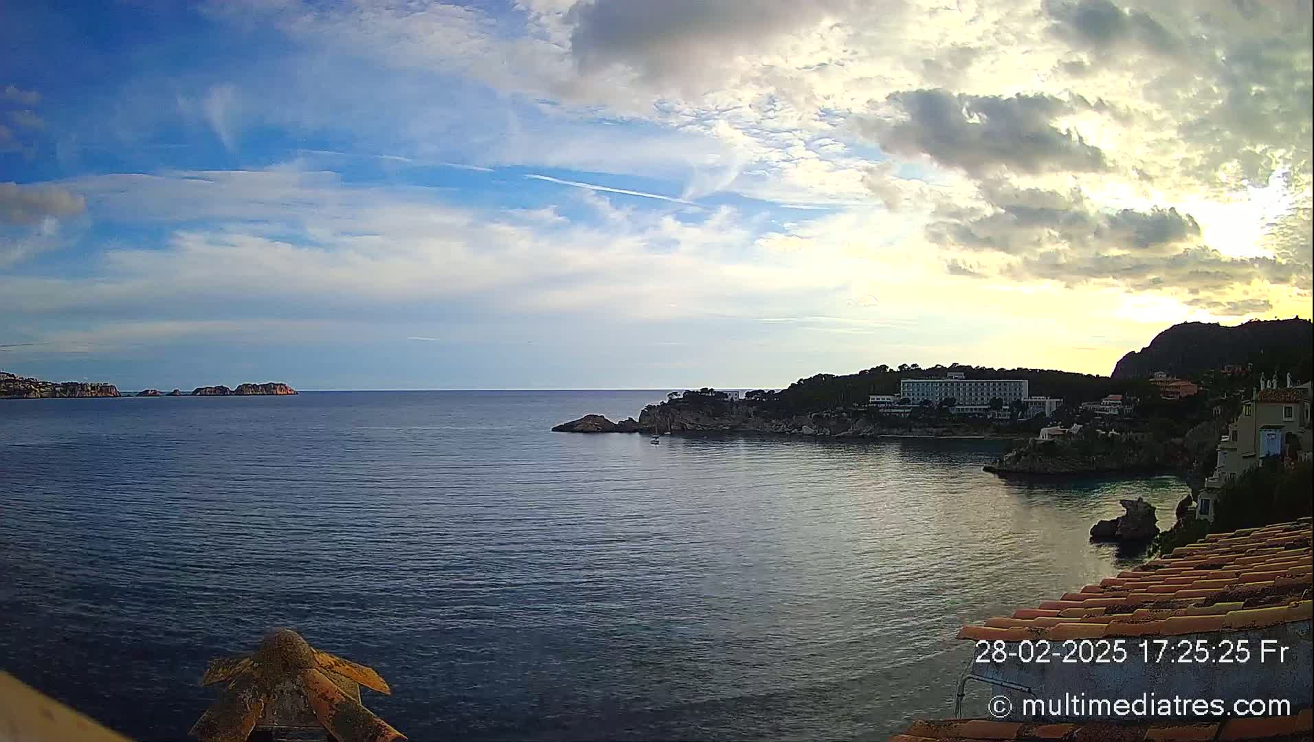 A partly cloudy sunset over a calm sea with rocky coastlines, including a large building situated on a cliff.