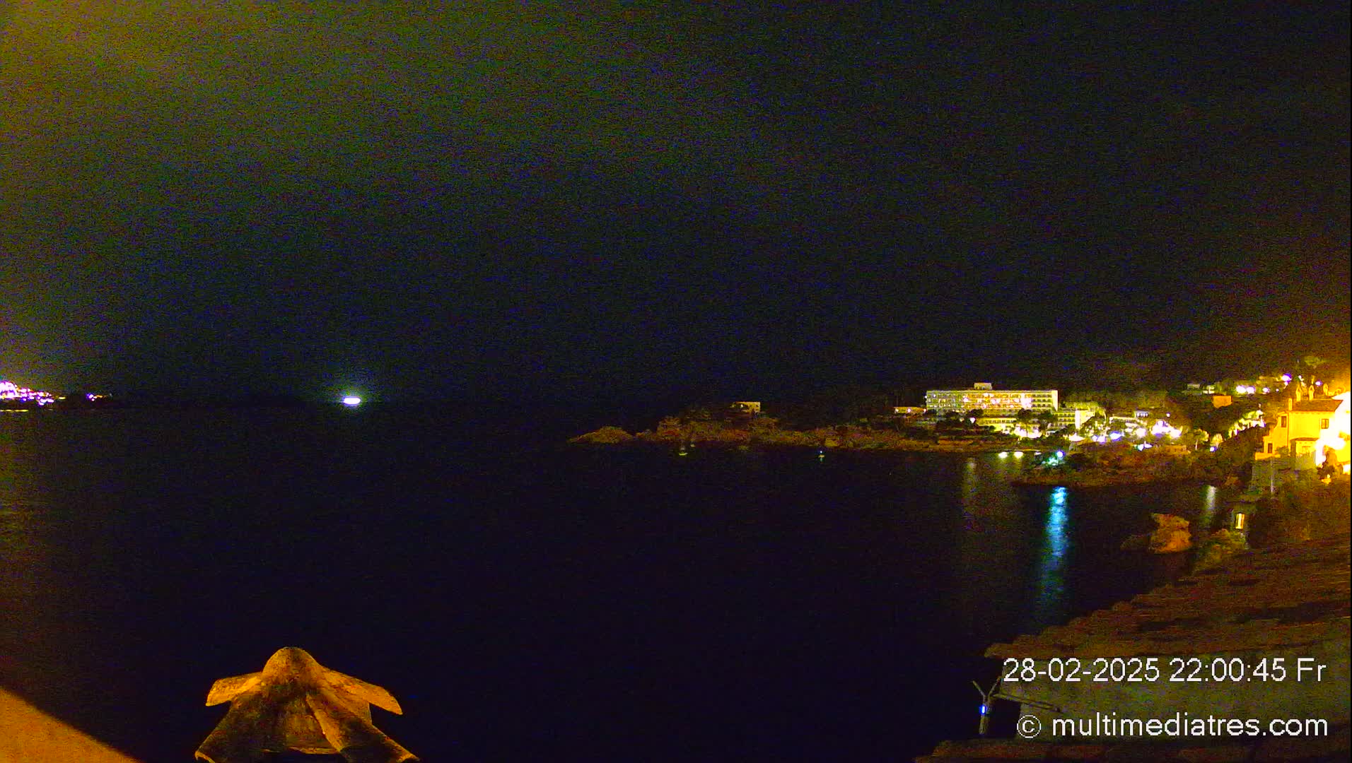 A nighttime view of a calm body of water with illuminated buildings on the opposite shore under a dark sky.