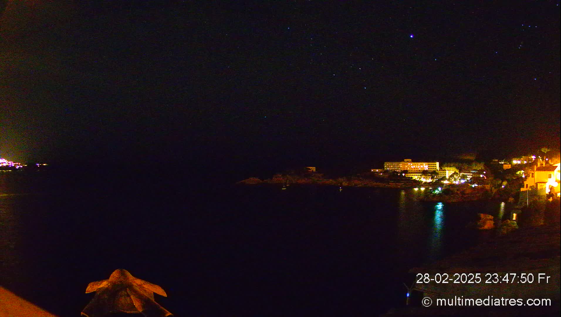 A nighttime view of a dark body of water with a starlit sky above and illuminated buildings along the shoreline.