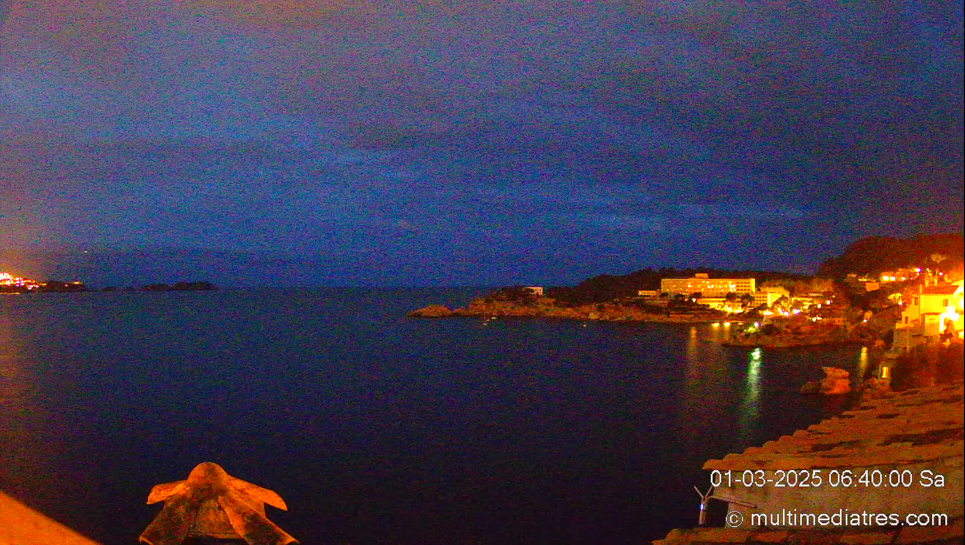 A nighttime coastal scene shows a dark body of water with illuminated buildings on the shore under a dark blue sky.