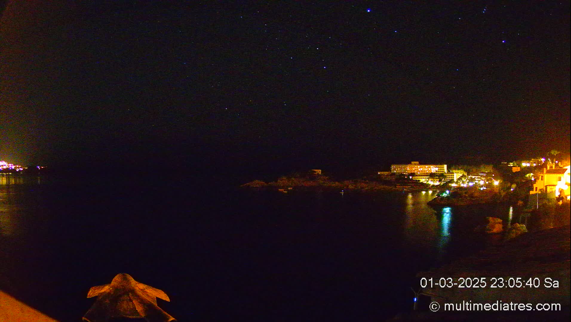 A nighttime view of a calm body of water with a town's lights illuminated on a hill across the water under a clear, starry sky.
