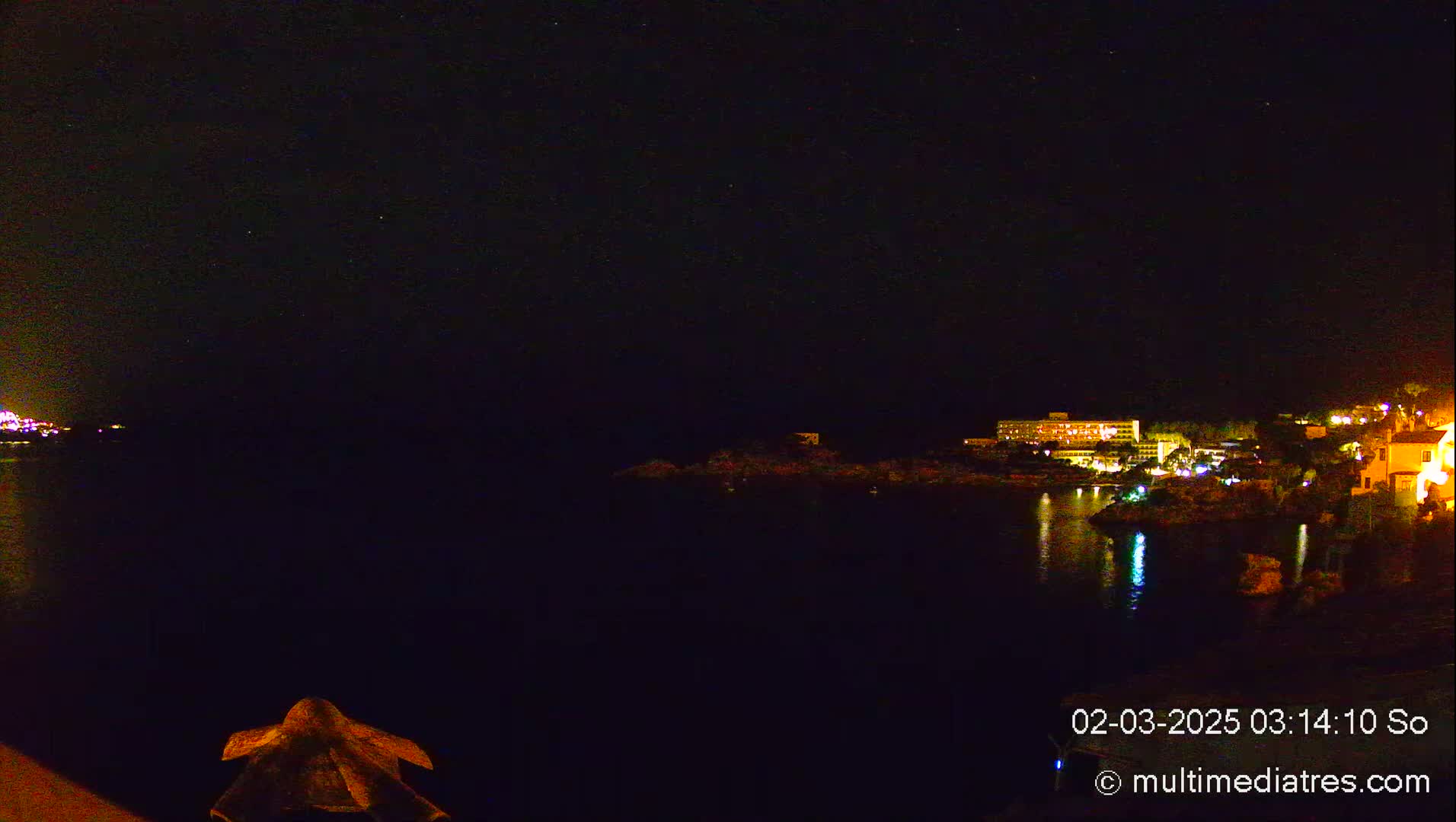 A nighttime view of a calm body of water reflecting the lights of buildings on the opposite shore under a clear, starry sky.