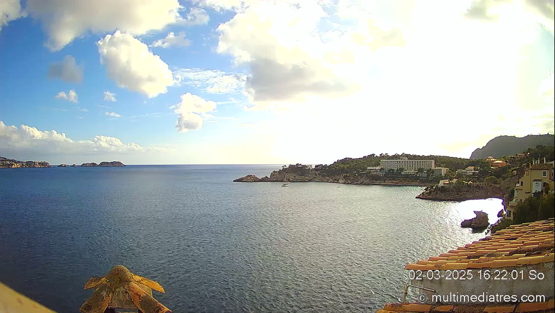A partly cloudy day shows a view of a calm ocean bay with a coastal town and small islands in the distance.