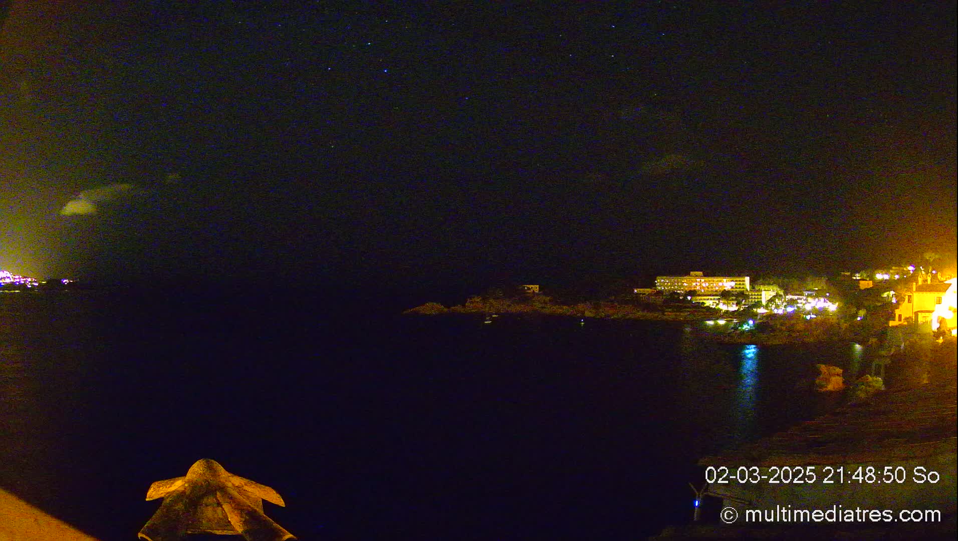 A nighttime coastal scene shows a dark body of water reflecting the lights of buildings on the opposite shore under a partly cloudy, starry sky.