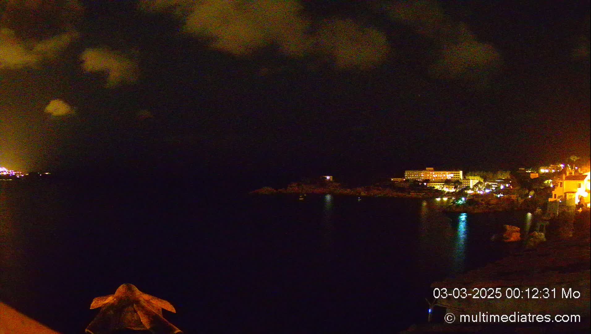 A nighttime view of a dark body of water with a shoreline of illuminated buildings under a partly cloudy sky.