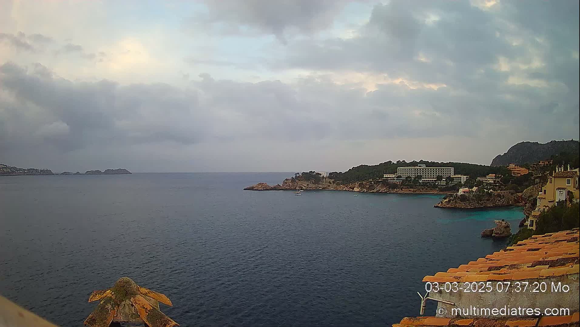 A coastal view shows a calm, dark-blue sea under a mostly cloudy sky, with a hotel and other buildings situated on a rocky shoreline.