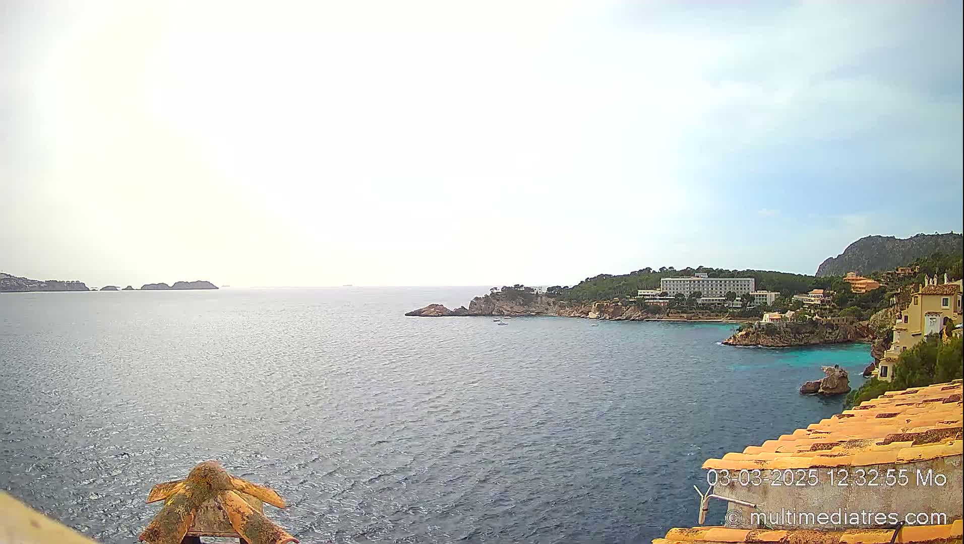 A calm, overcast day reveals a coastal scene with a hotel and buildings nestled along a rocky shoreline, overlooking a tranquil bay dotted with small islands.